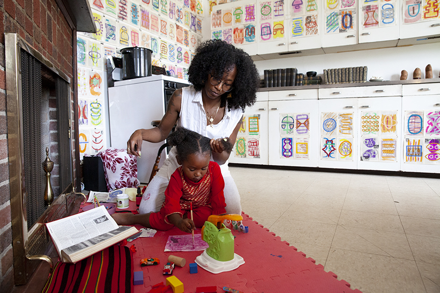 From Rites of Passage: 20/20 Vision. A black mother styles her daughters hair as she paints next to a fireplace in the room called "the soul kitchen."